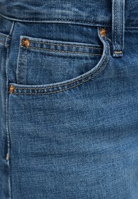 Gros plan sur un jean en denim bleu montrant la poche avant, la petite poche pour monnaie, la passants de ceinture et les rivets en cuivre avec des détails de couture visibles.