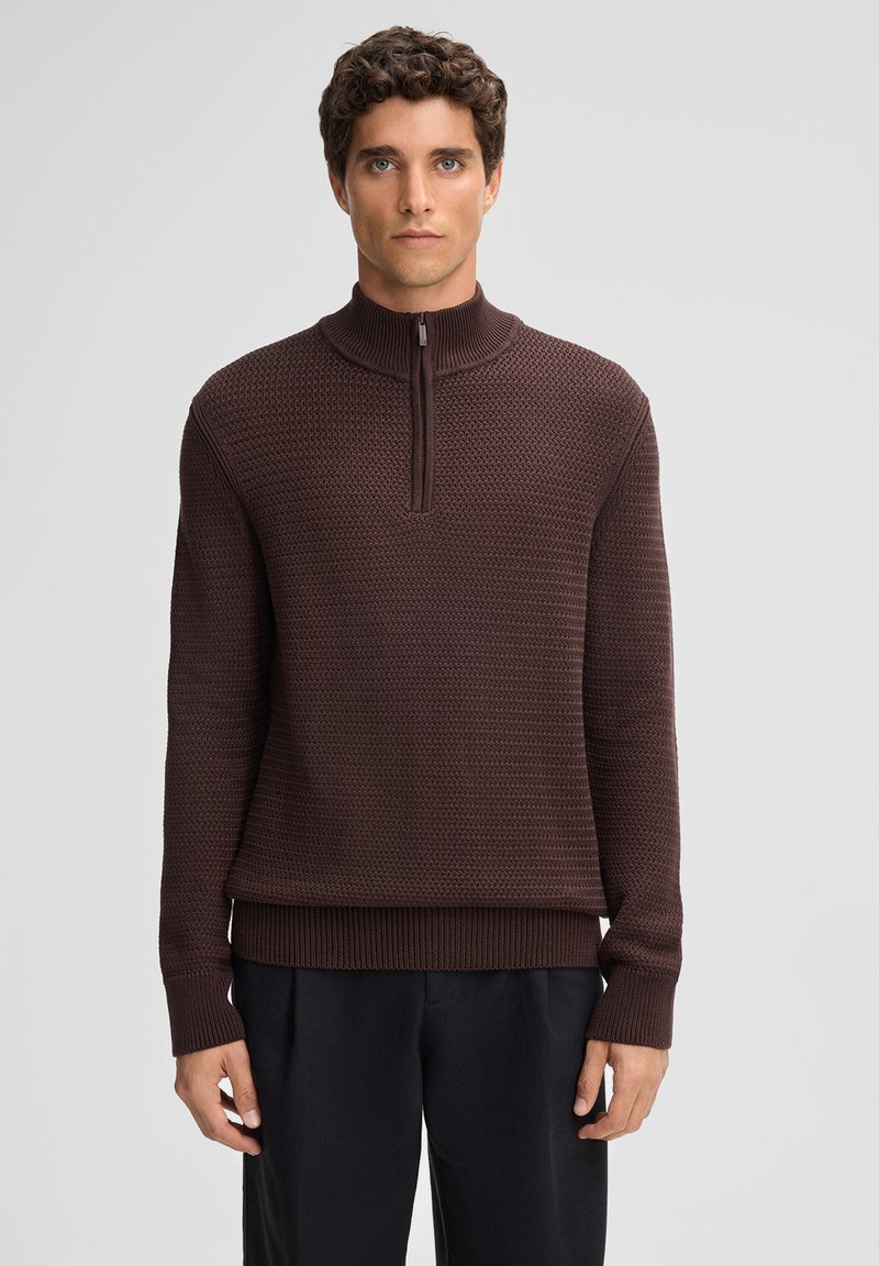 Brauner Strickpullover mit hohem Kragen, ausgestattet mit einem teilweisen Reißverschluss und geripptem Saum. Geprägtes Muster im gesamten Stoff.