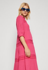 Vestido rosa de botão com mangas de três quartos, apresentando costura texturizada e um design em camadas. Usado com óculos de sol em formato de gato azuis.
