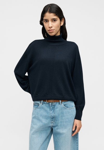 Ζιβάγκο πουλόβερ σε σκούρο μπλε ναυτικό χρώμα με άνετη εφαρμογή, ριμπ λεπτομέρειες και μακριά μανίκια, συνδυασμένο με ανοιχτό μπλε τζιν παντελόνι και καφέ ζώνη.