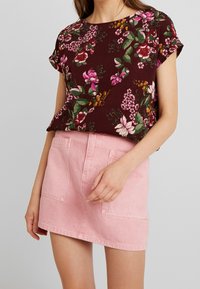 Top floreale a maniche corte in un intenso bordeaux scuro con fiori verdi, rosa e gialli, abbinato a una mini gonna in denim rosa chiaro con tasche frontali.