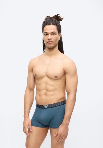 Άνδρας χωρίς πουκάμισο με κότσους dreadlocks, φοράει στενά μπλε εσώρουχα μπόξερ με μαύρη μέση, στέκεται μπροστά σε απλό λευκό φόντο.