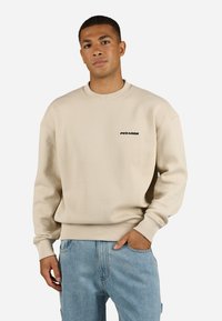 Beige textured sweatshirt with a crew neck and ribbed cuffs. Features a small black logo on the chest. Worn with blue jeans.