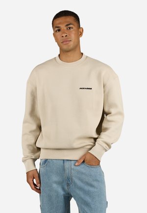 Beige textured sweatshirt with a crew neck and ribbed cuffs. Features a small black logo on the chest. Worn with blue jeans.