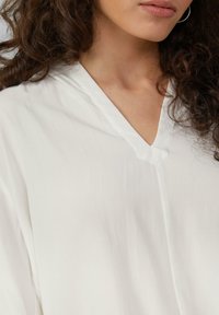 Biała bluzka z dekoltem w kształcie litery V, wykonana z miękkiego, gładkiego materiału, z subtelną teksturą i luźnym krojem, podkreślona widocznymi szwami.