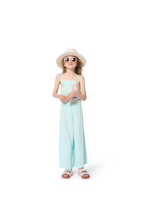 Jeune fille en tenue bleu clair avec des motifs d'étoiles de mer, portant un chapeau à large bord, des lunettes de soleil rondes et des sandales blanches, debout sur un fond blanc.