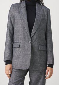 Blazer gris en tissu texturé, fermeture à un bouton, revers crantés et poches plaquées. Les poignets présentent un détail contrastant noir.