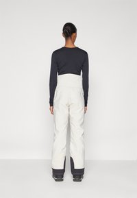 Weiße Schneehosen mit hoher Taille, elastischem Rücken und schmal zulaufenden Beinen. Schwarze Akzente an den Bündchen. Kombiniert mit einem langärmeligen, dunklen Oberteil.