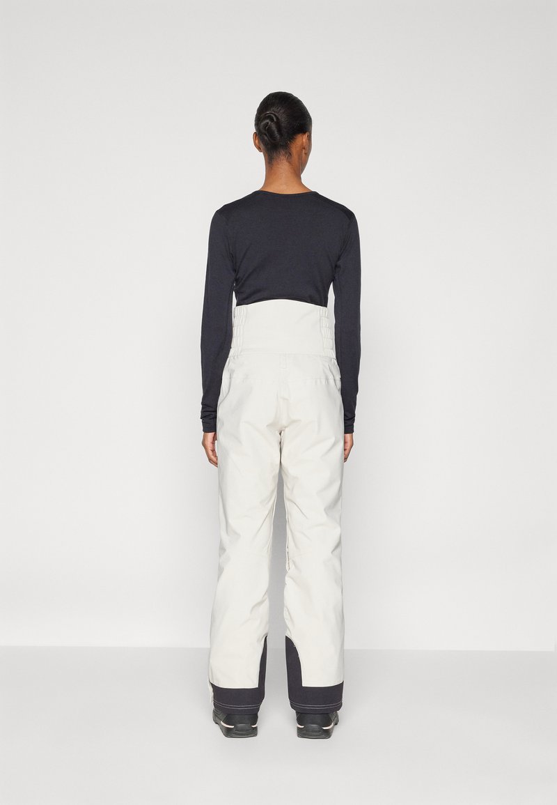 Białe spodnie narciarskie z wysokim stanem, elastycznym tyłem i zwężanymi nogawkami. Czarne akcenty przy mankietach. W komplecie z długim, ciemnym topem.