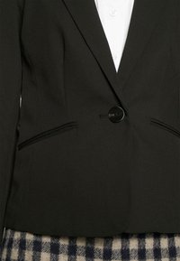 Blazer noir à un bouton porté sur une chemise blanche, associé à une jupe à carreaux noir et blanc.