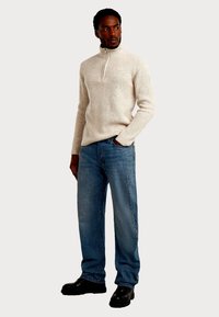 Beiger Rippenstrick-Pullover mit Reißverschluss, blaue Jeans mit geradem Bein und schwarze klobige Schuhe. Charakteristische Textur und lockere Passform.