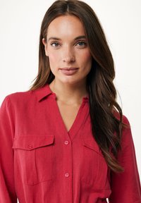 Röd linneskjorta med krage, två bröstfickor och knappr detaljer. Tyget har en strukturerad yta. Håret är långt och brunt.