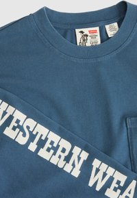 Blaues Baumwollhemd mit langen Ärmeln, das "WESTERN WEAR" in großen weißen Buchstaben zeigt, Rundhalsausschnitt und einer Brusttasche. Das Etikett enthält Markeninformationen.