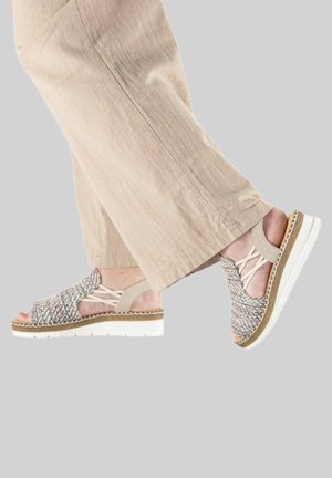 Pieds portant un pantalon beige à jambes larges et des sandales tissées multicolores à bout ouvert avec semelle blanche, avançant sur un fond gris.