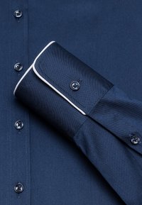Chemise bleu marine avec une texture lisse, des finitions blanches contrastantes sur les poignets et des boutons foncés le long de la patte de boutonnage.