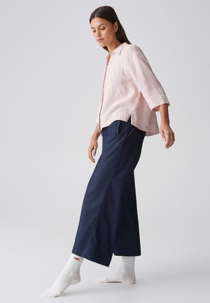 Frau trägt hellrosa Hemd und weite dunkelblaue Hose, balanciert auf einem Fuß vor schlichtem hellem Hintergrund.