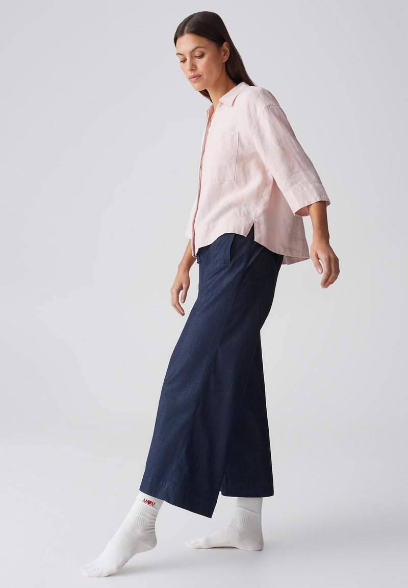 Woman wearing light pink shirt and wide dark blue pants, balancing on one foot against plain light background.