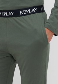 Maslinasto zeleni loungewear s crnim pojasom, s bijelim brendiranjem "REPLAY". Glatka tekstura s bočnim džepovima i opuštenim fitom.