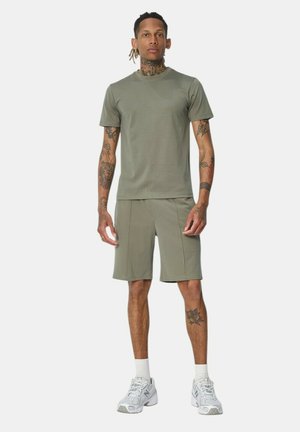 Homme tatoué debout portant une chemise à manches courtes vert olive et un short assorti avec des chaussettes blanches et des baskets grises sur fond blanc.