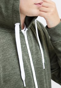 Groene rits hoodie van zacht materiaal, met een witte rits en trekkoorden, gestructureerde stof en een capuchon. Close-up van het nekgebied.