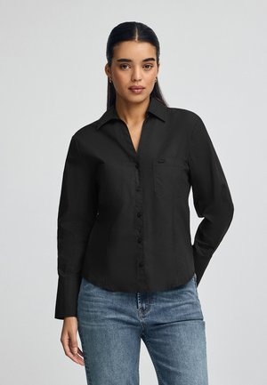Chemise noire à manches longues avec un col, dotée d'une poche poitrine unique et d'un tissu lisse, associée à un jean bleu clair.
