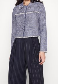 Blå tweedjacka med en kortad design, krage, knäppning med knappar och fransade kanter. Bärs över mörka byxor med ränder.