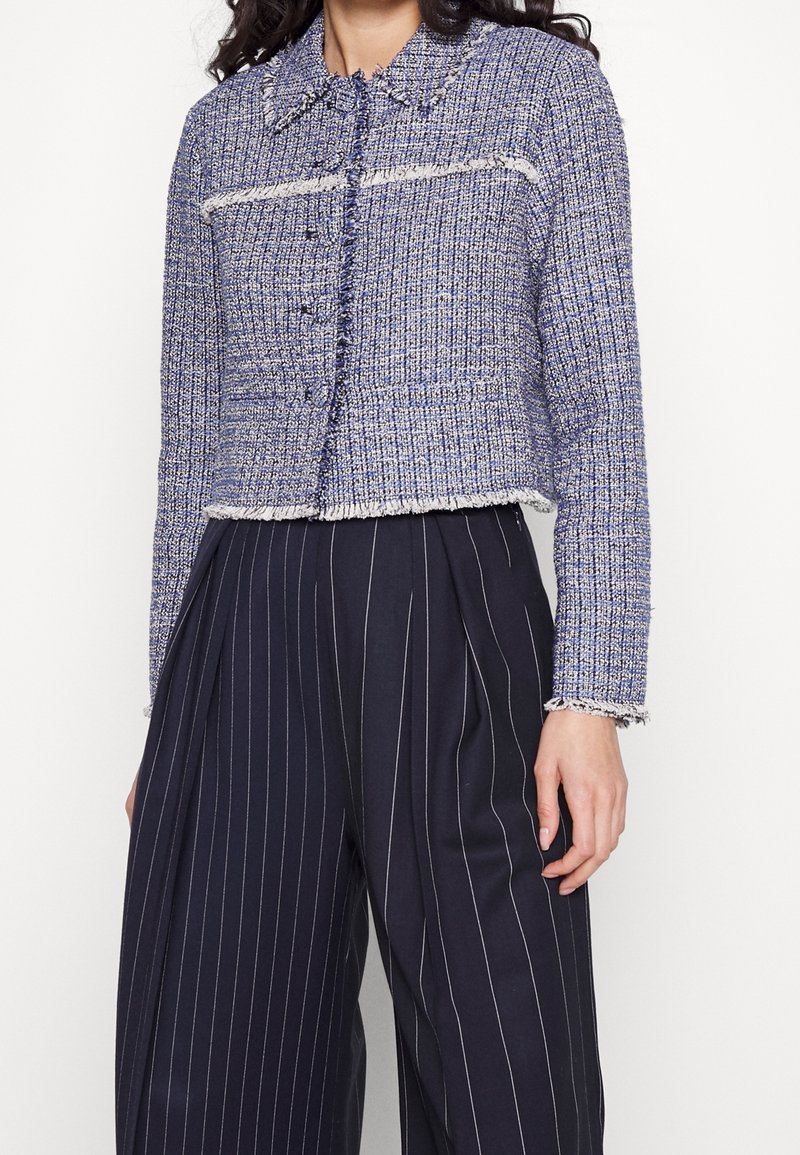 Blå tweedjacka med en kortad design, krage, knäppning med knappar och fransade kanter. Bärs över mörka byxor med ränder.