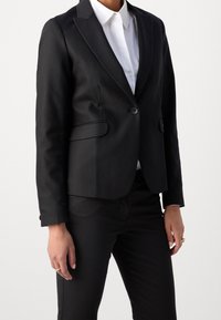 Blazer negro entallado con solapas de muesca, cierre de un solo botón y bolsillos frontales. Llevado sobre una camisa blanca con cuello, combinado con pantalones negros.