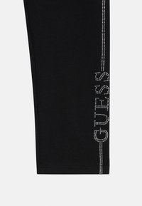 Schwarze Leggings in schmaler Passform mit einer vertikalen Linie von Strasssteinen, die "GUESS" entlang der Außennaht buchstabiert. Glatte Textur, dehnbares Material.