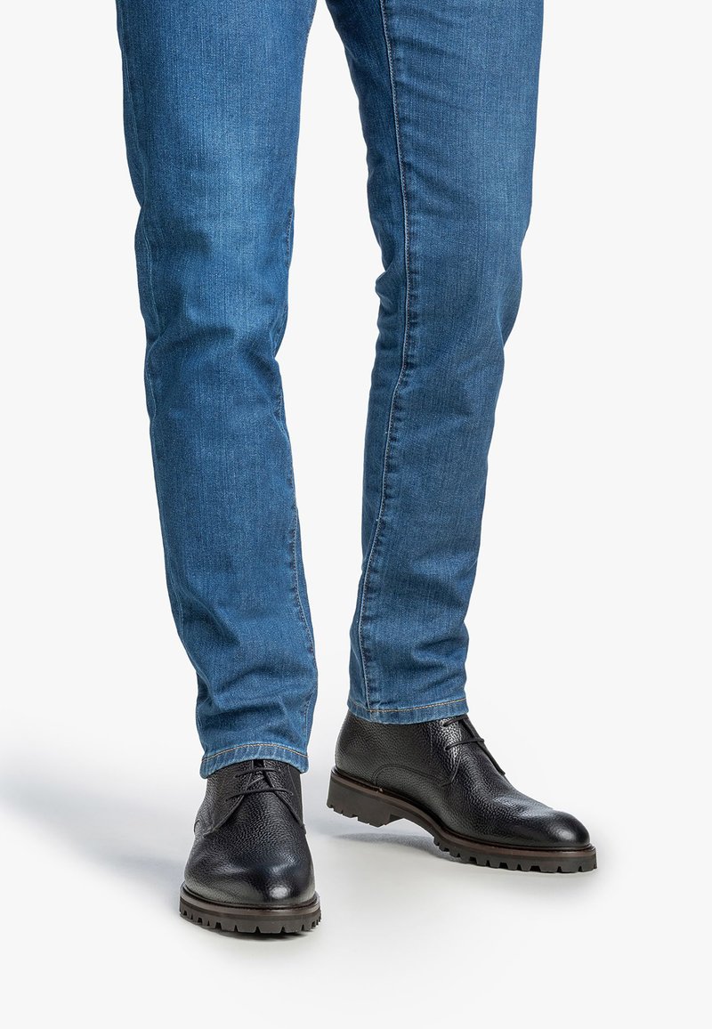 Zapatos de cuero negro con superficie texturizada y cordones, combinados con unos vaqueros ajustados azules. Los zapatos presentan una suela resistente y un diseño elegante.