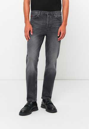 Man wearing faded black straight-leg jeans and black leather slip-on shoes, standing against a white background.