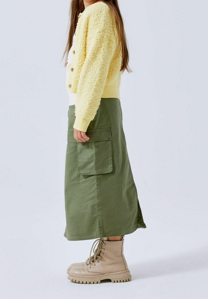 Fille debout de profil portant un cardigan jaune duveteux, une longue jupe verte olive avec une grande poche, et des bottes beige à lacets.