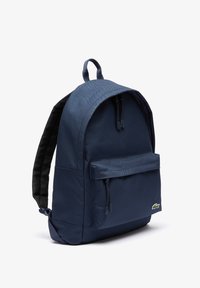 Sac à dos en tissu bleu marine avec un dessus arrondi, poche zippée à l'avant et bretelles réglables ; il possède un logo sur le bas à l'avant.
