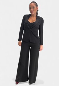 Completo nero con blazer su misura e pantaloni a gamba larga. Il tessuto ha una texture liscia, completata da décolleté rosse a punta.