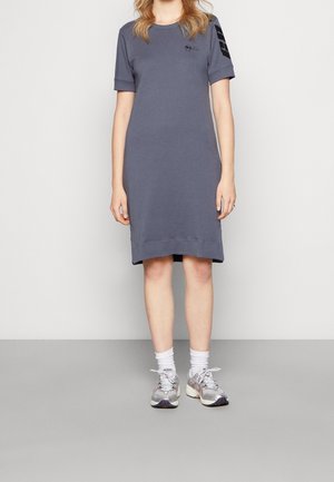 Jeune femme portant une robe grise décontractée à manches courtes, des chaussettes blanches et des chaussures de sport argentées, debout devant un mur uni.
