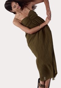 Olivegrünes Maxi-Kleid mit plissierter Textur, dünnen Trägern und gerafftem Oberteil. Getragen mit schwarzen Sandalen mit einem Nietenriemen.