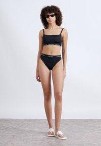 Σετ μαύρο μαγιό μπικίνι με τοπ μπραλέ και ψηλόμεσο σλιπ με λογότυπο Calvin Klein. Το μοντέλο φοράει γυαλιά ηλίου και λευκά σανδάλια slide.