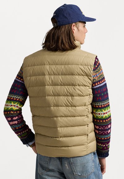 Beige puffer vest with horizontal quilted pattern, worn over a multicolored patterned knitted sweater, complementing denim jeans and a cap.