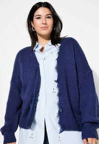 Navyblauw gebreid vest met een gestructureerde afwerking, open voorkant, lange mouwen en rafelige randen; gecombineerd met een lichtblauw overhemd met knopen.