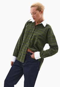 Camicia a quadri verdi in flanella con colletto e polsini bianchi, dotata di una vestibilità ampia e chiusura a bottoni. Indossata con pantaloni in denim scuri.