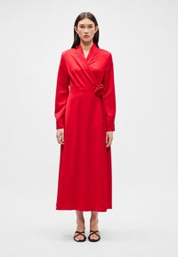 Czerwona satynowa sukienka typu wrap z długimi rękawami, akcentem w postaci róży w talii oraz długością do podłogi, charakteryzująca się gładką, błyszczącą fakturą.