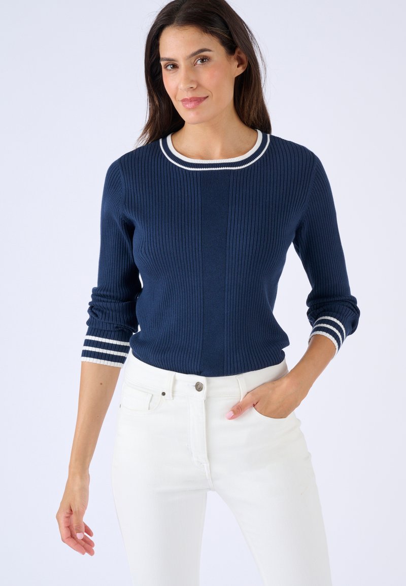 Mujer vistiendo un suéter de canalé azul marino con rayas blancas en el cuello y los puños, combinado con pantalones blancos, posando con una mano en el bolsillo.