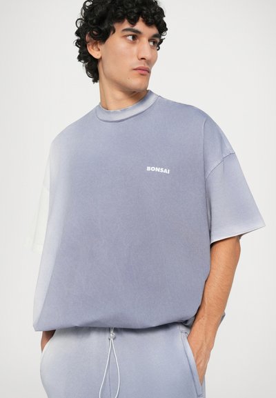 Tricou oversized de culoare albastru deschis, cu mâneci scurte, având un decolteu rotund și un mic logo alb "BONSAI" pe față.