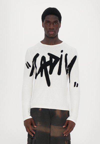 Homme portant une chemise blanche à manches longues avec un grand texte manuscrit noir "HAPILY" et un pantalon cargo noir avec des taches brunes, se tenant devant un fond blanc.