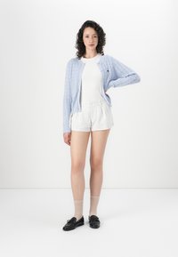 Light blue knitted cardigan, white fitted top, striped shorts, and black loafers. Medium-length curly hair, standing pose on a white background.