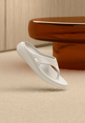 Enkele witte Lacoste teenslipper die zweeft boven een beige tapijt met op de achtergrond een bruin transparant rond tafeltje.