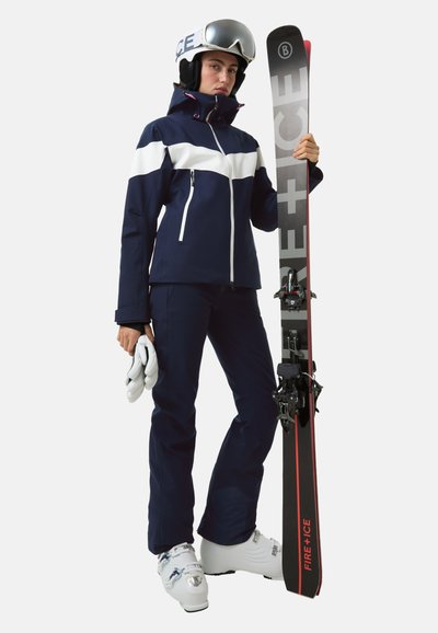 Strój narciarski składający się z granatowej kurtki z białymi akcentami, granatowych spodni oraz białych butów narciarskich. W rękach czarne i czerwone narty. Do kompletu kask i rękawice.