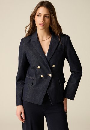 Kobieta w ciemnoniebieskiej dwurzędowej marynarce z złotymi guzikami, w czarnych spodniach, stoi na beżowym tle.