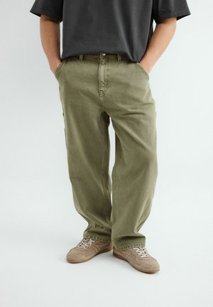 Homme portant un pantalon ample vert olive, des baskets beiges et un t-shirt gris foncé, les mains dans les poches, debout sur un fond blanc uni.