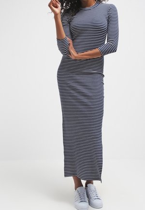 Kobieta w granatowo-białej sukience maxi w paski z długim rękawem i białych sneakersach, stojąca na jednolitym tle.